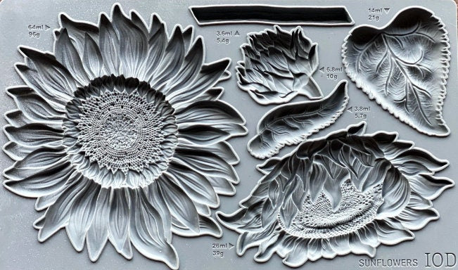 May include: A silicone mold for baking or crafting. The mold features a large sunflower design with a detailed center and petals. There are also smaller sunflower designs, leaves, and bud shapes. The mold is gray and has measurements and weights printed on it. The text on the mold reads 'SUNFLOWERS IOD'.
